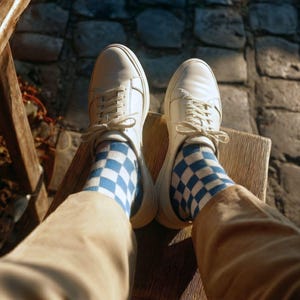 May include: A pair of white sneakers with matching laces, worn with blue and white checkered socks. The shoes are resting on a wooden surface, with a cobblestone background visible.