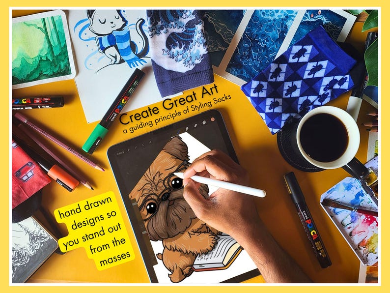 Overhead view of an artist's desk showing the creative process for Styling Socks. An iPad displays a hand-drawn Griffon dog illustration, surrounded by Posca markers, colored pencils, and watercolor paintings, emphasizing unique, hand-drawn designs.