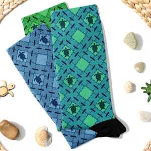 Könnte beinhalten: Drei Paar Socken mit einem geometrischen Muster aus blauen und grünen Quadraten und einem Meeresschildkröten-Design. Die Socken sind gefaltet und auf einem weißen Hintergrund angeordnet.