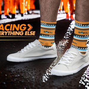 May include: A pair of white sneakers with orange and blue striped socks featuring a racing car design. The socks have the text "RACING > EVERYTHING ELSE" on them.