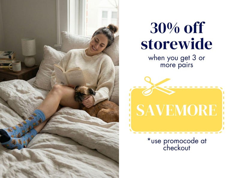 A promotional graphic showing a woman cozy in bed reading a book, wearing the blue and yellow Griffon reading socks next to a real Brussels Griffon dog. Text indicates a 30% off storewide sale when buying 3 or more pairs using code SAVEMORE.