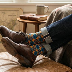 Puede incluir: Zapatos de cuero marrón estilo brogue con calcetines estampados en tonos azules, naranjas y amarillos. Los calcetines son visibles por encima de los zapatos, y la persona lleva vaqueros azules con los puños doblados. La escena sugiere relajación.