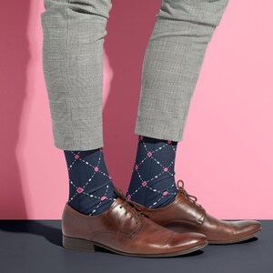 May include: A pair of brown leather dress shoes with navy blue argyle socks featuring white and pink diamond patterns. The socks are worn with gray plaid trousers. The background is a pink wall and a dark blue floor.