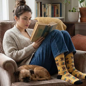 A cozy lifestyle photograph of a woman sitting in an armchair, engrossed in a hardcover book. She is wearing the yellow Griffon reading socks, and a real Brussels Griffon dog is sleeping curled up beside her.