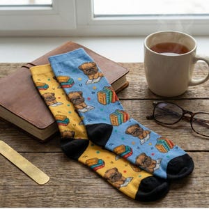 Mismatched blue and yellow crew socks featuring illustrations of Griffon Bruxellois dogs reading books. The socks rest on a rustic wooden table next to a steaming mug, reading glasses, and a leather journal. A cozy book lover gift scene.