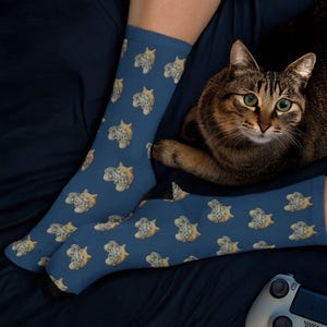 A lifestyle photo of a person wearing the blue variation of the Novelty Gaming Cat Themed Socks snuggled up in bed, with their cat and game controller