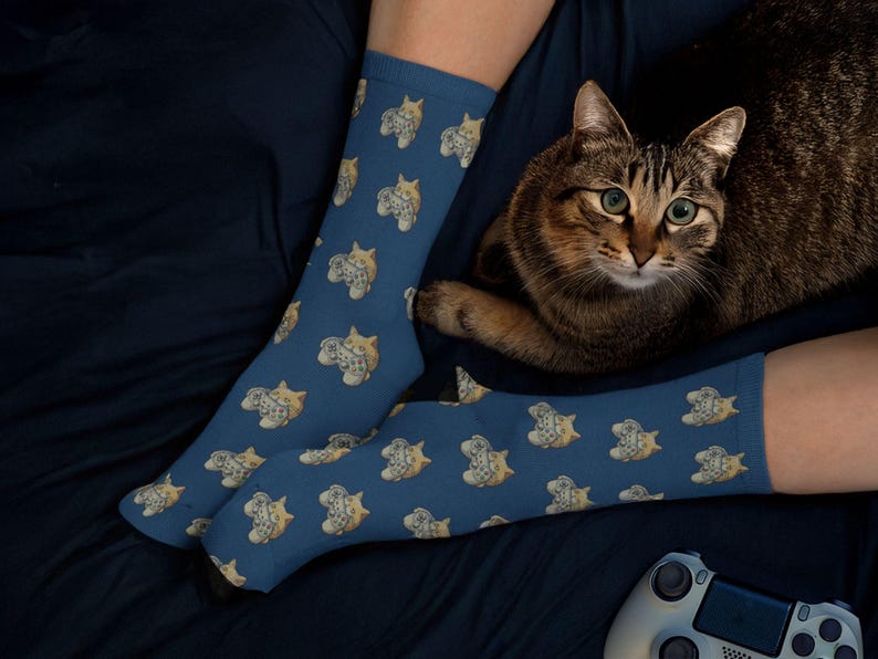 A lifestyle photo of a person wearing the blue variation of the Novelty Gaming Cat Themed Socks snuggled up in bed, with their cat and game controller