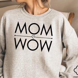 May include: Light grey sweatshirt with the words "MOM WOW" in bold black letters. The word "MOM" is above the word "WOW", separated by a horizontal line with a heart in the middle. The sweatshirt is a casual style.