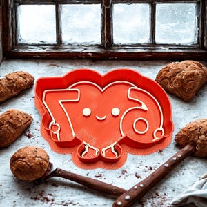 May include: A red cookie cutter in the shape of the number 76 inside a heart, with a smiling face. The text "CUSTOM SIZES AVALIBLE" is above the cookie cutter. The cutter is surrounded by baked goods and wooden tools.