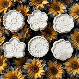May include: Six white, flower-shaped cookie cutters in various designs are arranged on a background of yellow and brown sunflowers. The cutters include a daisy, a rose, and other floral shapes, perfect for baking.