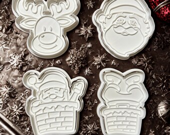May include: Four white Christmas-themed cookie cutters. Designs include a reindeer, Santa Claus face, Santa Claus in a chimney, and a chimney with a Santa Claus hat. The cutters are on a dark, textured surface with snowflake and ornament details.