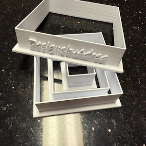 May include: Stack of white geometric cookie cutters in various sizes. The largest cutter is rectangular, with the text "DesignsbyAdrea" embossed on its side. The other cutters are square and rectangular.