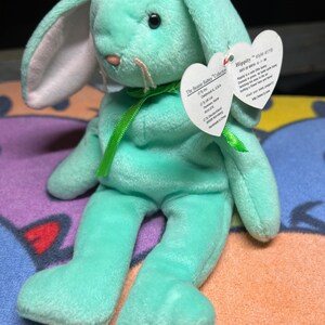Może przedstawiać: Zielony pluszowy królik Beanie Baby o imieniu Hippity z zieloną wstążką wokół szyi. Etykieta zawiera napis "Hippity" i "Data urodzenia: 8-1-97".