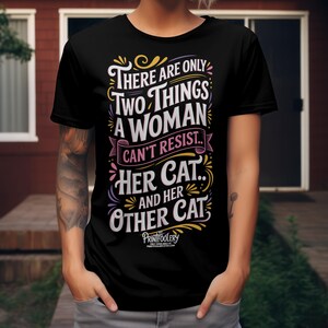 A person with tattooed arms wearing a black t-shirt featuring a cat lover graphic.