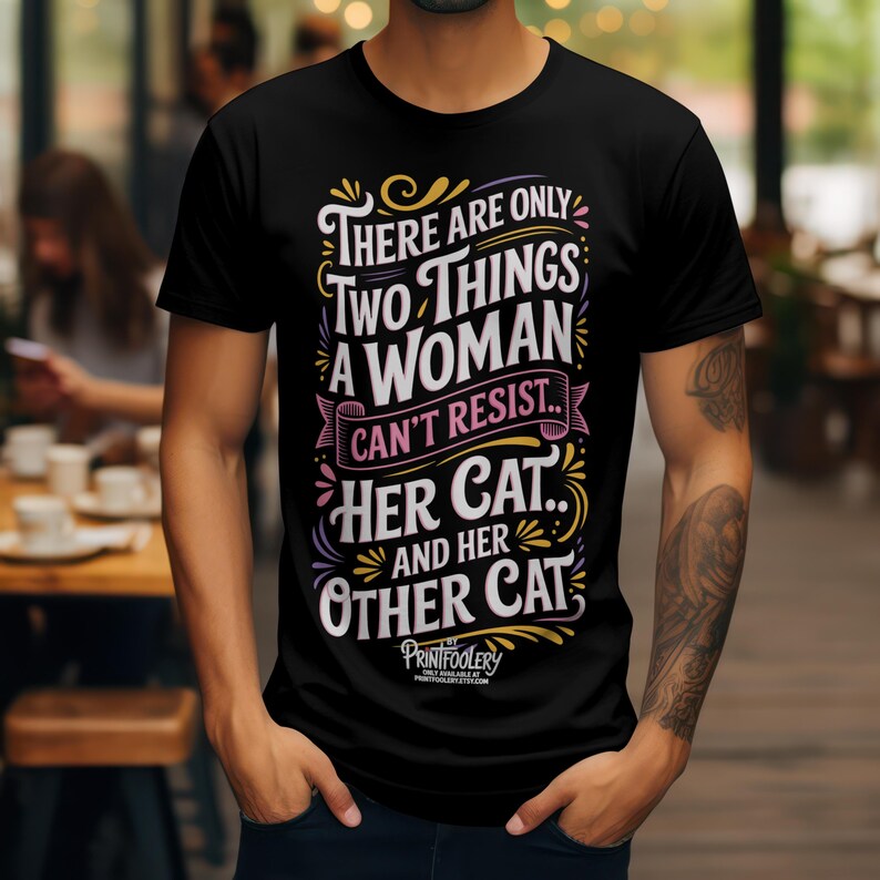 A man wearing a black t-shirt with a humorous cat graphic standing in front of a cafe interior.