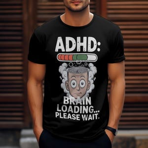Peut inclure: T-shirt noir avec le texte "ADHD:" au-dessus d'une barre de chargement. En dessous, une tête de dessin animé avec un cerveau et les mots "BRAIN LOADING... PLEASE WAIT." Le t-shirt est à manches courtes.