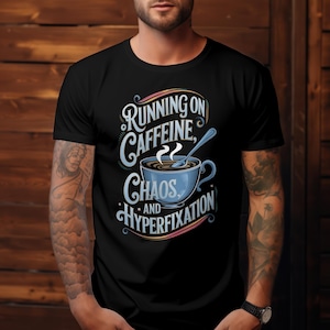 Op de afbeelding: Zwart T-shirt met een afbeelding van een koffiekopje en de tekst "Running on Caffeine, Chaos, and Hyperfixation".