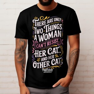 A tattooed man wearing a black t-shirt with colourful cat lover typography against a white brick wall.