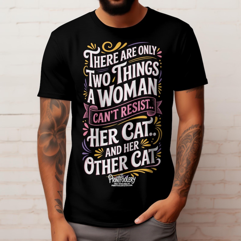 A tattooed man wearing a black t-shirt with colourful cat lover typography against a white brick wall.
