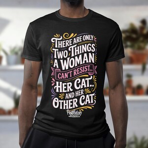 A person wearing a black t-shirt with cat typography standing in a room with potted plants on shelves.