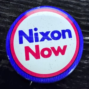 May include: A round button with a blue border and the words "Nixon Now" in blue and red lettering. The button has a white background with a red ring around the text.