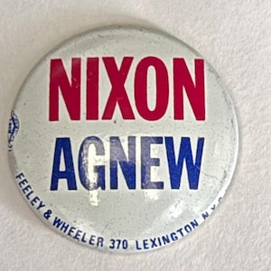 May include: A round, vintage political campaign button with the words "NIXON AGNEW" in red and blue block letters. The button has a light gray background and the text "FEELEY & WHEELER 370 LEXINGTON N.Y.C." is printed around the edge.