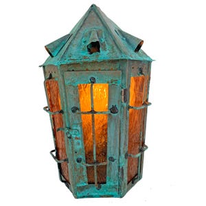 May include: A hexagonal lantern with a weathered teal and copper patina finish. The structure features a pointed roof, metal framing with horizontal bars, and textured amber-colored glass panels that emit a warm, glowing light from within.