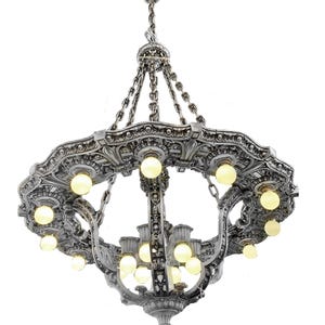 May include: Ornate silver chandelier with multiple light bulbs. The chandelier has a detailed, decorative design with a circular shape and is suspended from chains. The light bulbs are evenly spaced around the fixture.