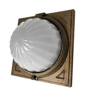 May include: A square wall-mounted light fixture featuring a weathered bronze-toned metal frame with triangular corner accents. A large, white, fluted glass dome shade is centered within the frame, secured by a hinged top and a small bottom latch.