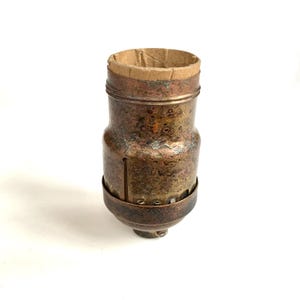 May include: A vintage brass lamp socket with a weathered, aged appearance. The socket has a cylindrical body with a textured surface and a brown paper insert at the top. A metal ring secures the base.