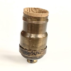 May include: Vintage brass light socket with a paper insert at the top. The socket has a cylindrical shape with a slightly wider base and a screw at the bottom. The metal has a tarnished, aged appearance.