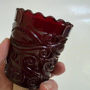 May include: A deep red glass vessel featuring a scalloped top rim and an intricate embossed pattern of swirling scrolls and beaded lines. The glass has a glossy, translucent finish that catches the light, highlighting the raised relief textures.