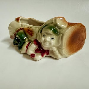 May include: A vintage ceramic planter shaped like a smiling elf emerging from a tree stump. The planter features a cream-colored base with green, red, and brown accents. The elf has a green hat and a red bow. The tree stump has a detailed wood grain design.