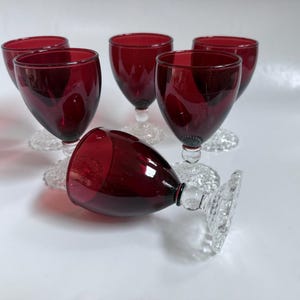 May include: A set of six wine glasses featuring deep red glass bowls and clear, textured glass stems and bases. Five of the glasses are standing upright, while one glass is positioned horizontally in the foreground, showcasing the hobnail pattern.