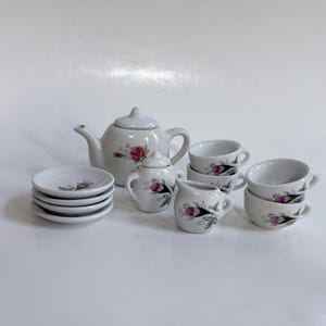 May include: A white porcelain tea set with pink rose floral designs. The set includes a teapot, sugar bowl, creamer, six saucers, and six teacups. The tea set is a miniature size.