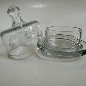 May include: A clear glass butter dish with a lid. The lid has a handle and a floral design etched on the side. The dish is open, revealing the base and the dish's interior.