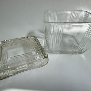 May include: A clear glass square container with a matching lid, featuring vertical ribbed detailing on the sides and a geometric grid pattern on the top surface of the lid. The glass is transparent and has a smooth, polished finish throughout.