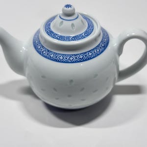 May include: A white porcelain teapot featuring a blue geometric pattern around the lid and neck. The body of the teapot is decorated with a subtle, embossed rice grain pattern, and it includes a matching lid with a small knob handle.
