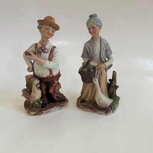 May include: Two ceramic figurines of an elderly couple. The man wears a hat, suspenders, and holds an object. A rooster stands at his feet. The woman holds a basket and stands near a goose. The figurines are painted in muted colors.