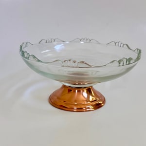 May include: A clear glass pedestal bowl with a scalloped rim and a copper-colored base. The bowl is designed to hold fruit or desserts. The copper base adds a touch of elegance to the design.