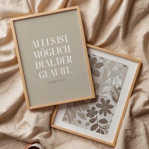 Könnte beinhalten: Zwei gerahmte Kunstdrucke mit Holzrahmen liegen auf einer beigen Oberfläche. Ein Druck zeigt den Text "ALLES IST MÖGLICH DEM, DER GLAUBT." in Weiß. Der andere zeigt ein Blumenmuster in Grau- und Weißtönen. Eine Hand hält eine weiße Tasse mit Kaffee.