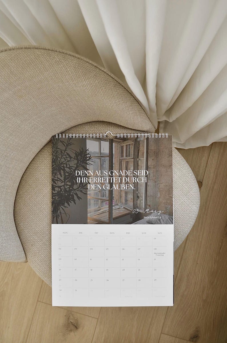 May include: A calendar with a photograph of a window and text in German, "DENN AUS GNADE SEID IHR ERRETTET DURCH DEN GLAUBEN." The calendar is open to March 3rd. The calendar is on a beige chair with a white curtain in the background.