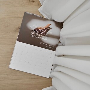 May include: A calendar with the text "JESUS SPRICHT KOMMT HER ZU MIR, ALLE, DIE IHR M&Uuml;HSELIG UND BELADEN SEID" is open on a wooden surface. White fabric is draped in the background. The date "11 November" is visible.