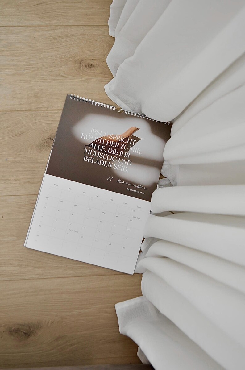 May include: A calendar with the text "JESUS SPRICHT KOMMT HER ZU MIR, ALLE, DIE IHR M&Uuml;HSELIG UND BELADEN SEID" is open on a wooden surface. White fabric is draped in the background. The date "11 November" is visible.