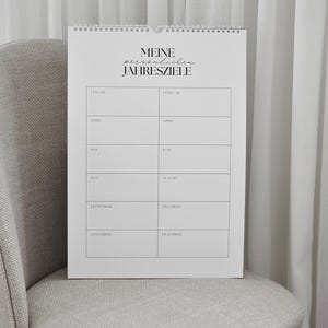 May include: A white spiral-bound calendar with the text "MEINE pers&ouml;nlichen JAHRESZIELE" at the top. The calendar is open to a page with monthly columns. The calendar is on a gray chair.