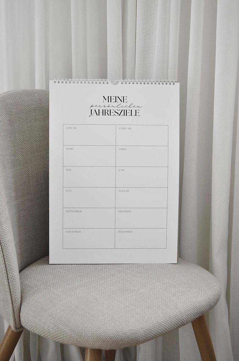 May include: A white spiral-bound calendar with the text "MEINE pers&ouml;nlichen JAHRESZIELE" at the top. The calendar is open to a page with monthly columns. The calendar is on a gray chair.