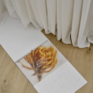 May include: A white spiral-bound calendar with a floral image and the text "ICH BIN BELEUCH ALLE TAGE." The calendar is open to a month view, with a wooden floor and white fabric in the background.