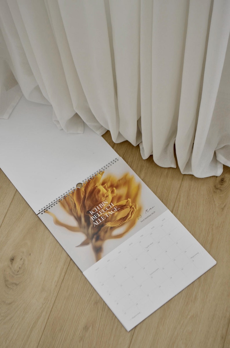 May include: A white spiral-bound calendar with a floral image and the text "ICH BIN BELEUCH ALLE TAGE." The calendar is open to a month view, with a wooden floor and white fabric in the background.