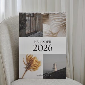 May include: A 2026 calendar with a spiral binding, featuring four images: a window scene, a close-up of a mushroom, a dried flower, and a lighthouse. The word "KALENDER" and the year "2026" are printed on the cover. The calendar is resting on a beige chair.