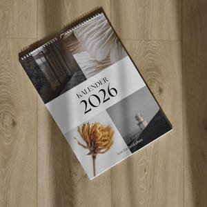 May include: A spiral-bound 2026 calendar with a collage of images, including a flower, architectural details, and a lighthouse. The word "KALENDER" and the year "2026" are prominently displayed. The calendar is lying on a wooden surface.
